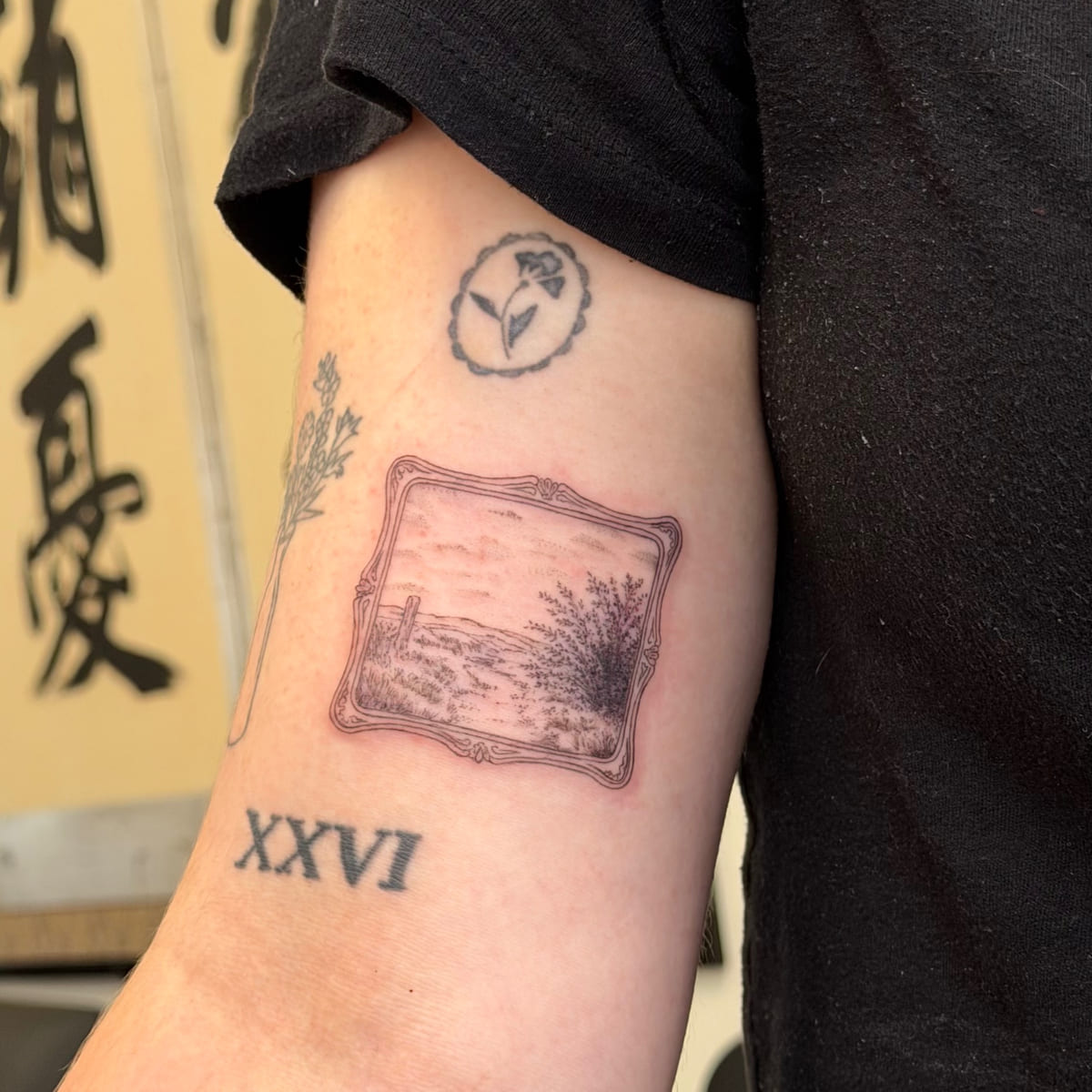 Landscape painting in an ornate frame with the numeral XXVI, a small flower in a ring above, fine line tattoo