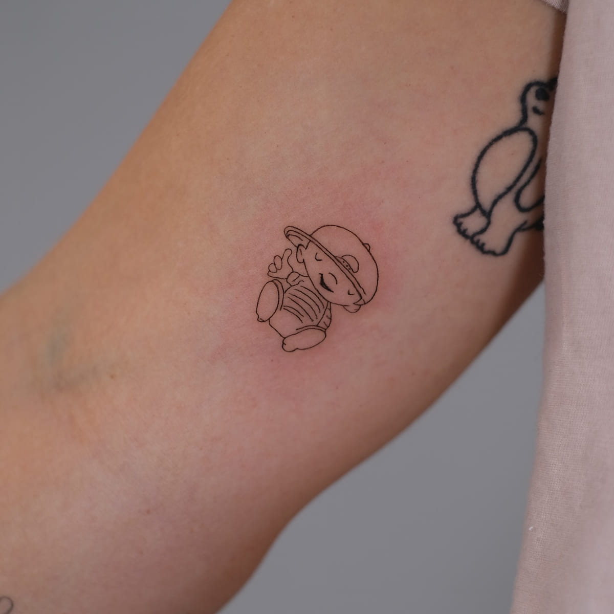 Small sitting character in a cap, fine line tattoo