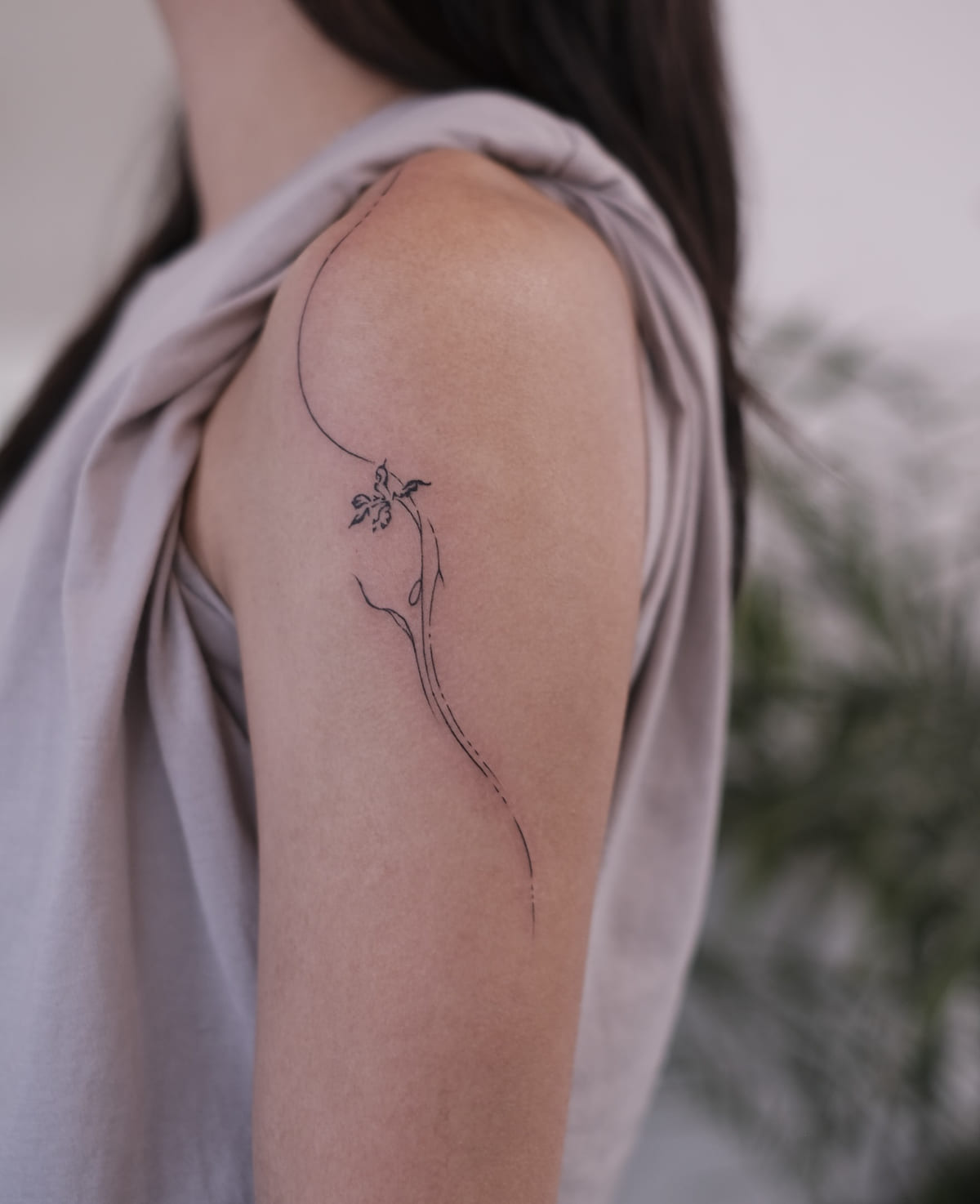 Long curving stem with a small flower, following the shoulder, fine line tattoo