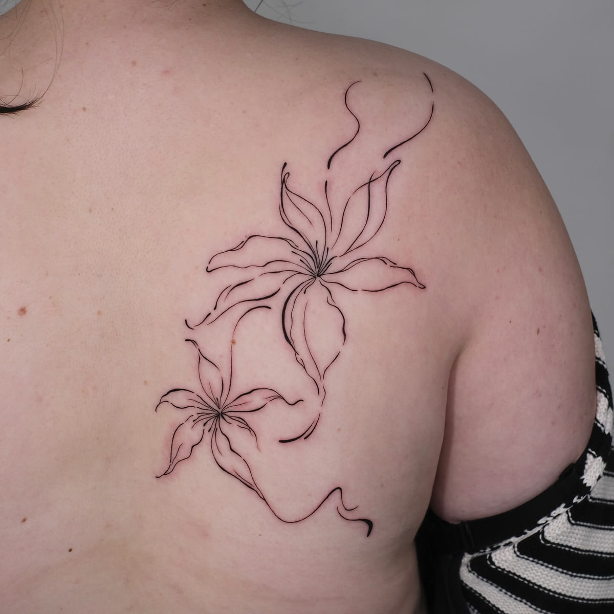 Two large lilies in flowing line across the shoulder blade, fine line tattoo