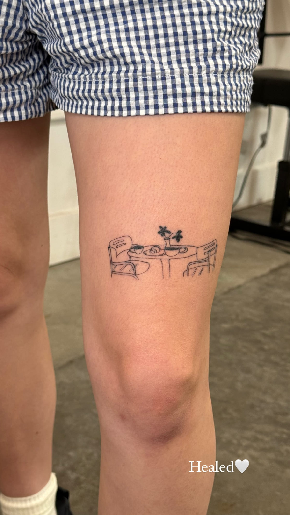 Small cafe table scene above a knee, healed fine line tattoo