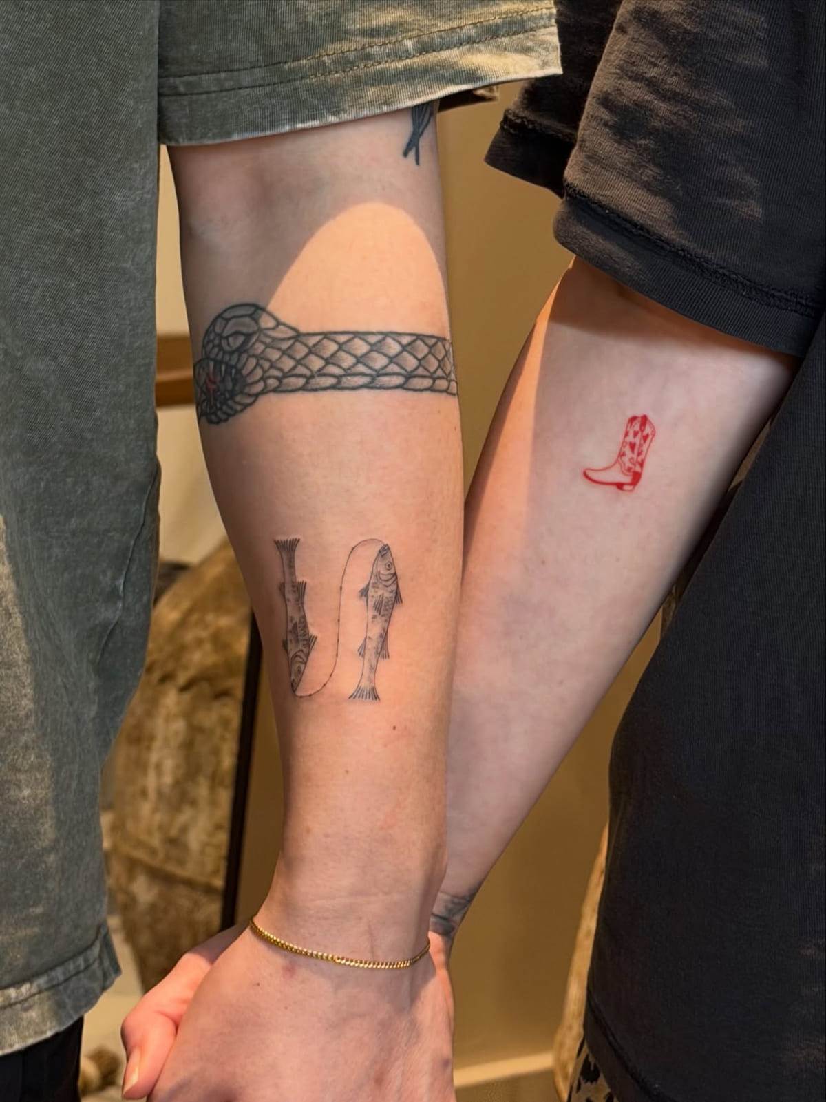 Couple holding hands: a red cowboy boot on one arm, two hanging fish on the other, fine line tattoo