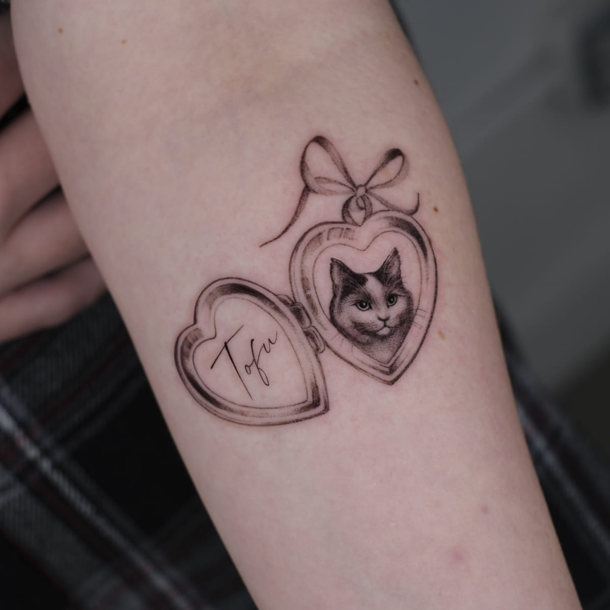 Open heart locket with a cat portrait and the name Tofu, bow on top, fine line tattoo