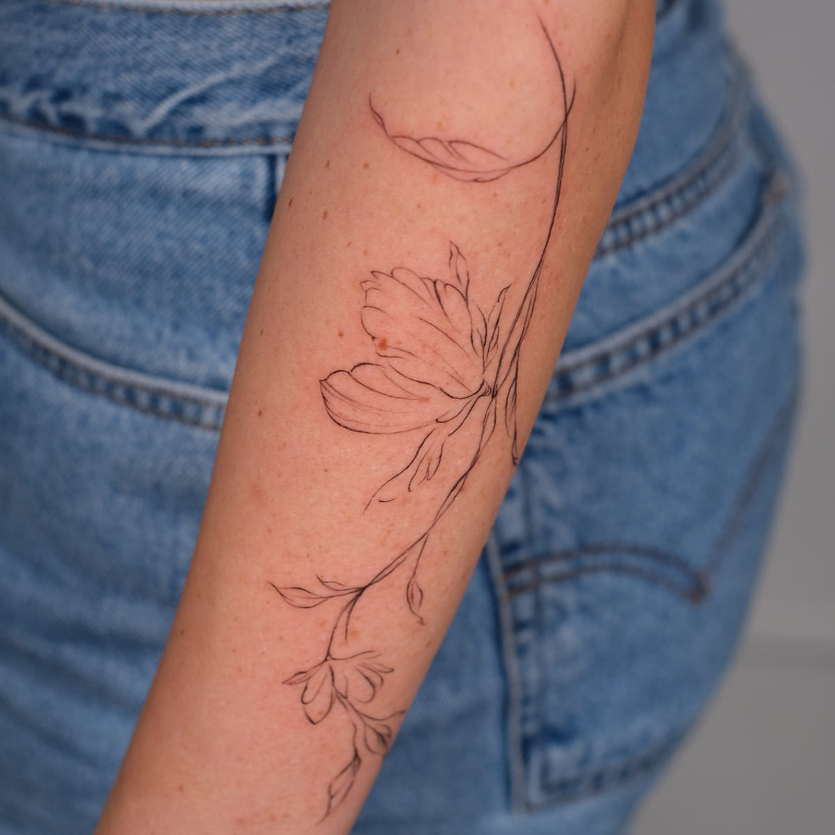 Large single flower on a long stem flowing down the forearm, fine line tattoo