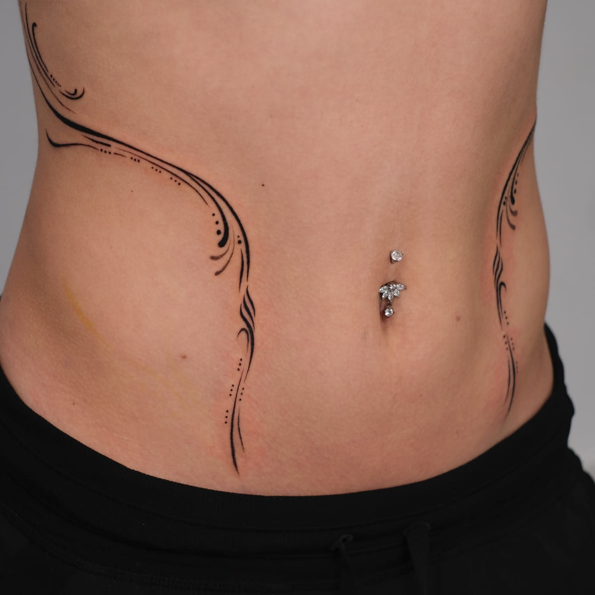 Ornamental flowing lines framing both sides of the abdomen, fine line tattoo