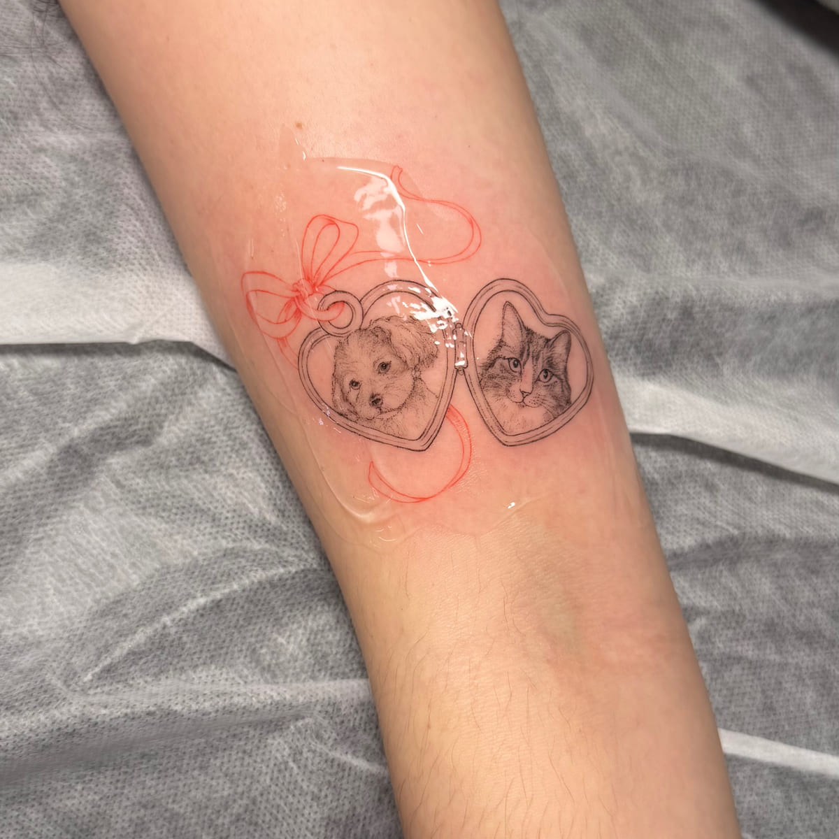 Two heart lockets tied with a red bow, a dog in one and a cat in the other, fine line tattoo
