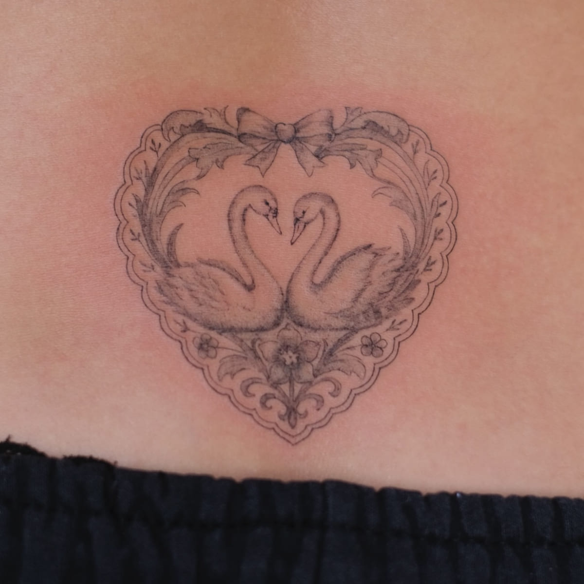 Two swans inside an ornate heart frame with a bow, fine line tattoo