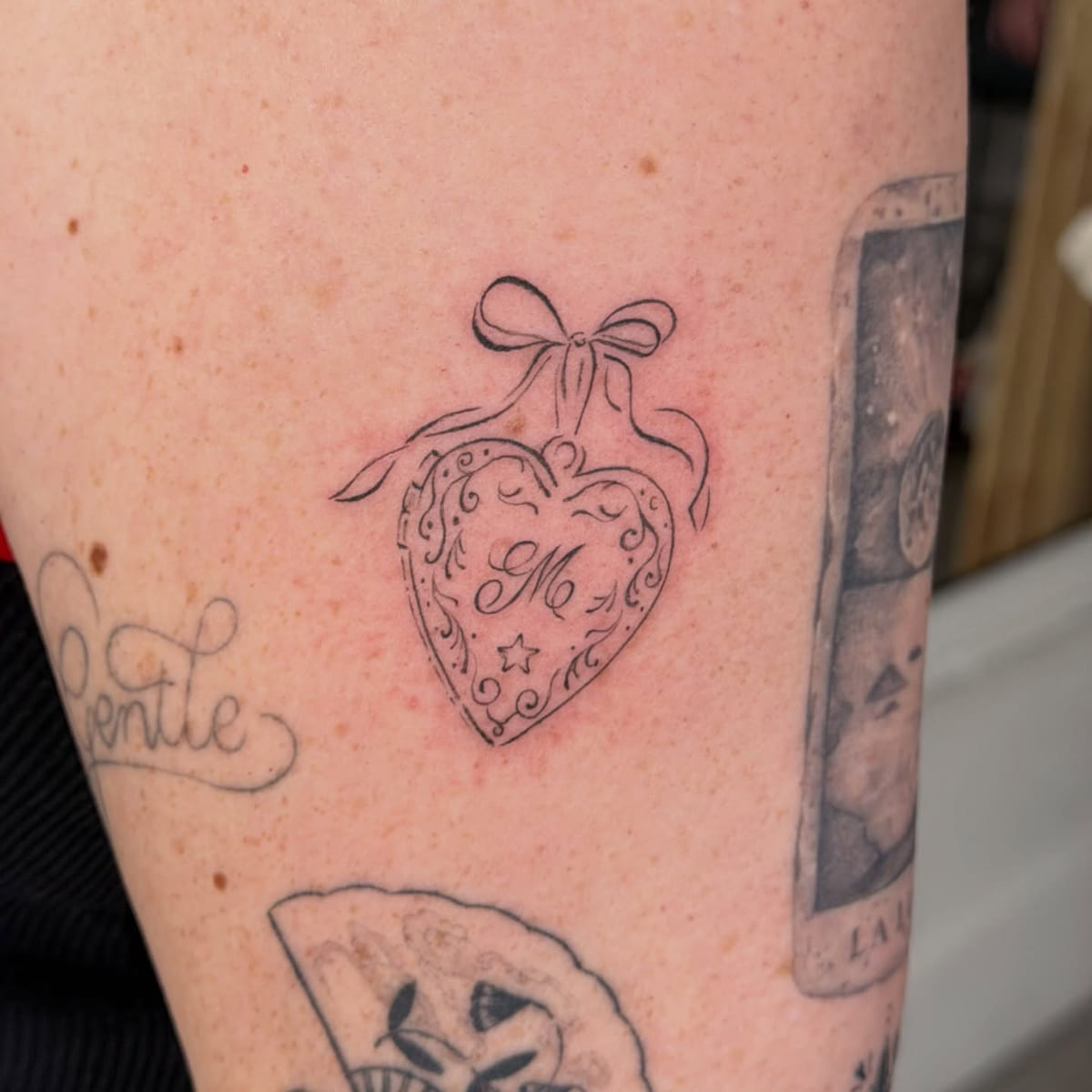 Heart locket with a bow and a script initial, fine line tattoo