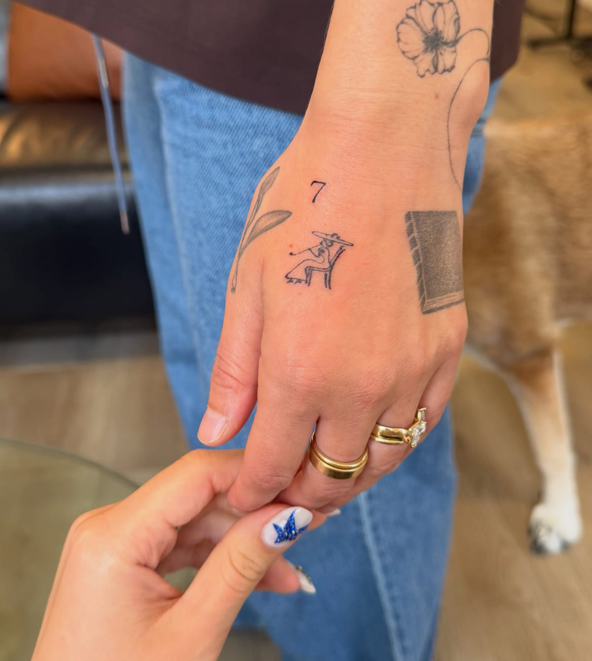 Tiny dancing figure with a 7 above, on the back of the hand, fine line tattoo