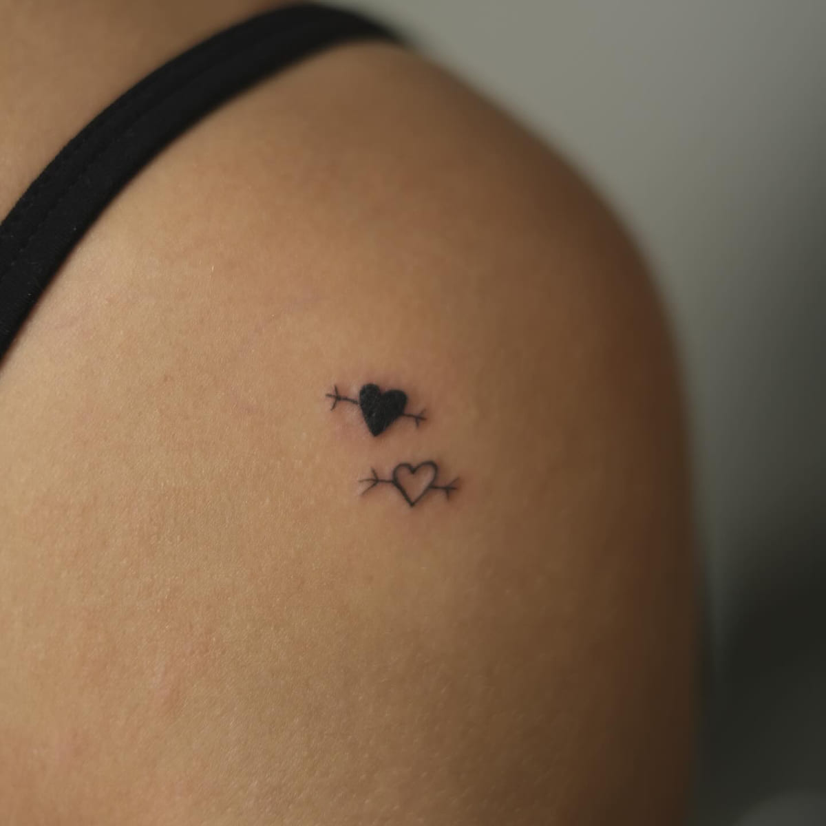 Two tiny hearts pierced by arrows, one solid and one outline, fine line tattoo